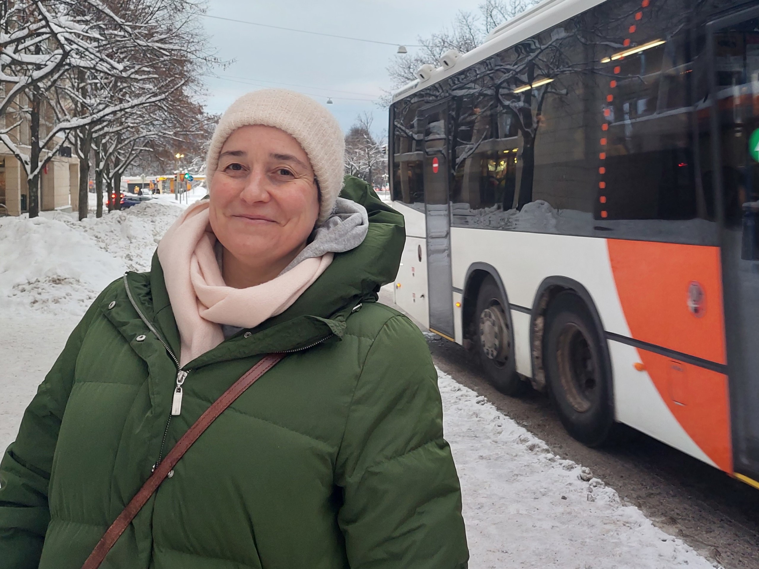 Sari Järvinen seisoo talvisella kadulla bussipysäkillä. Takana menee bussi.
