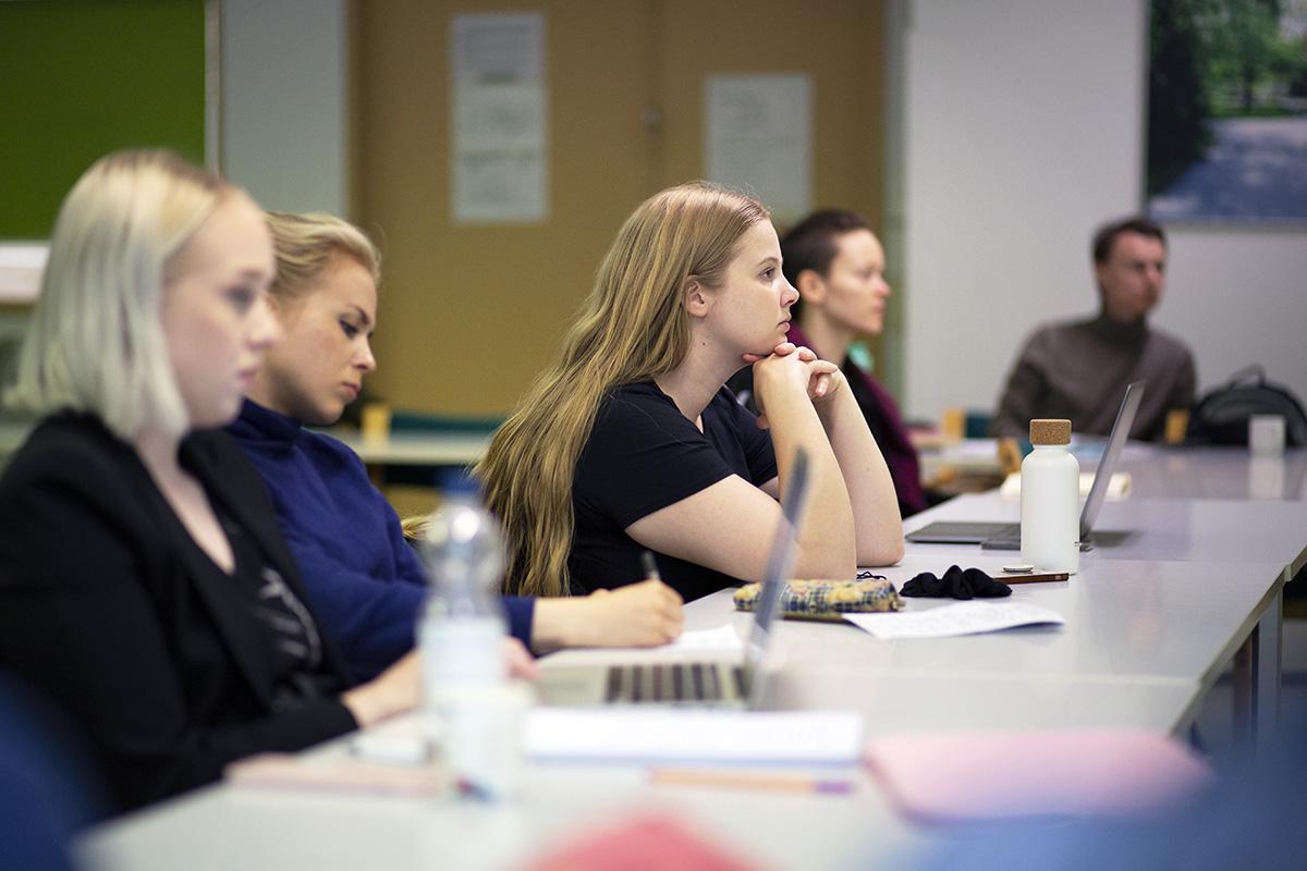 Nuoria pöydän ääressä kuuntelemassa opetusta. Pöydällä tietokoneita ja muistiinpanovälineitä.