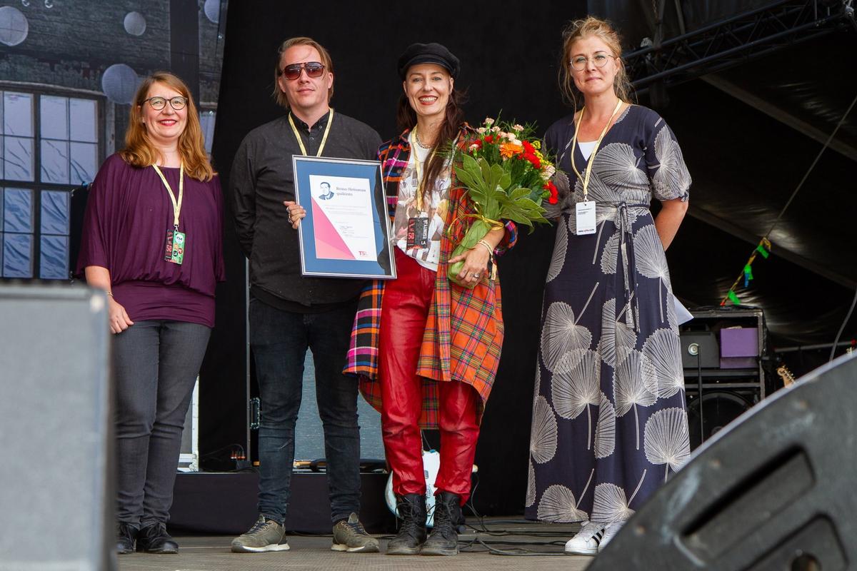 Mariska lavalla vastaanottamassa palkintoa, jota on luovuttamassa kolme henkilöä.