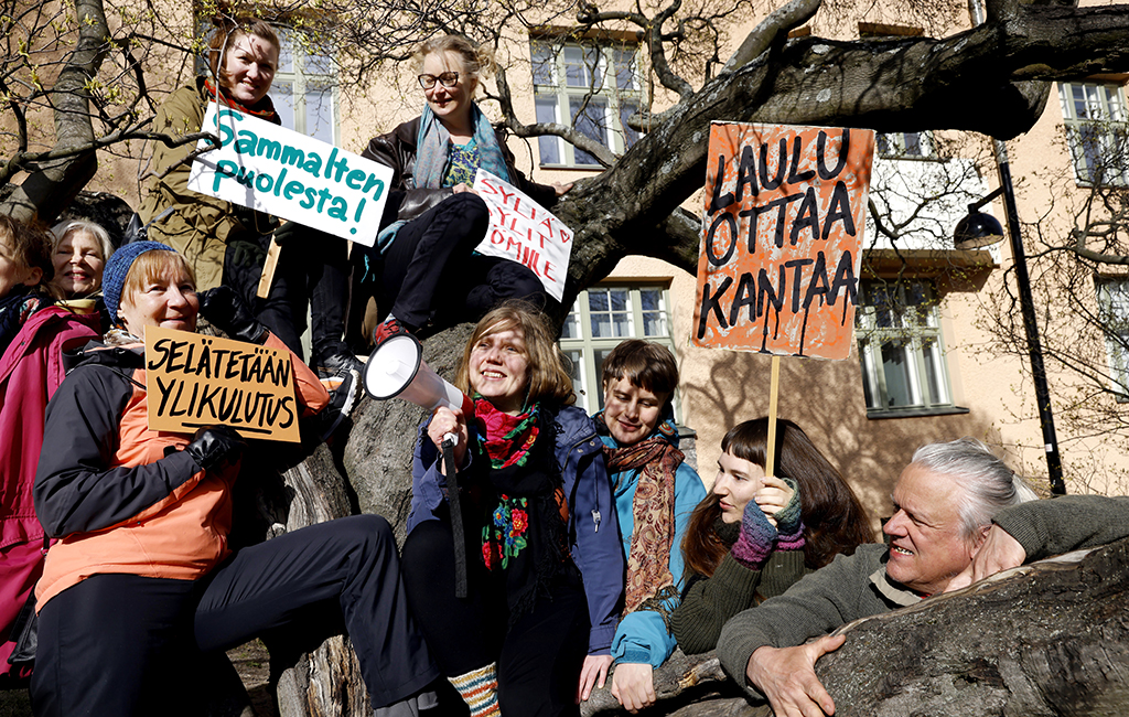 Laulu ottaa kantaa -kuorolaiset poseeraavat puun ympärillä, osa on kiivennyt puuhun. Mukana kylttejä, jossa lukee mm. ”Sammalten puolesta” ja ”Selätetään ylikulutus”.