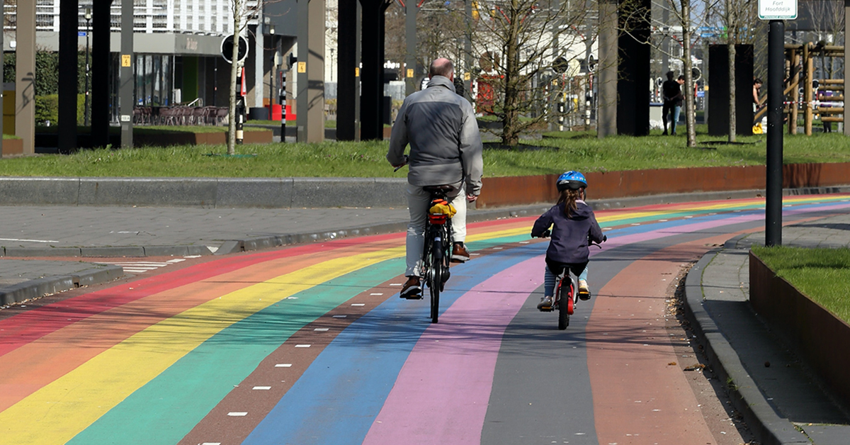 Isä ja lapsi pyöräilevät sateenkaaren värein maalatulla pyörätiellä.