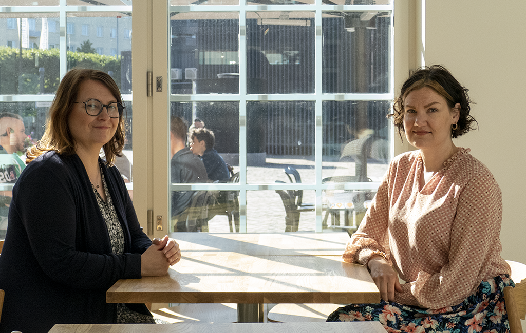 Säde Tahvanainen ja Pinja Kuusenaho istuvat pöydän ääressä ja katsovat hymyillen kameraan.