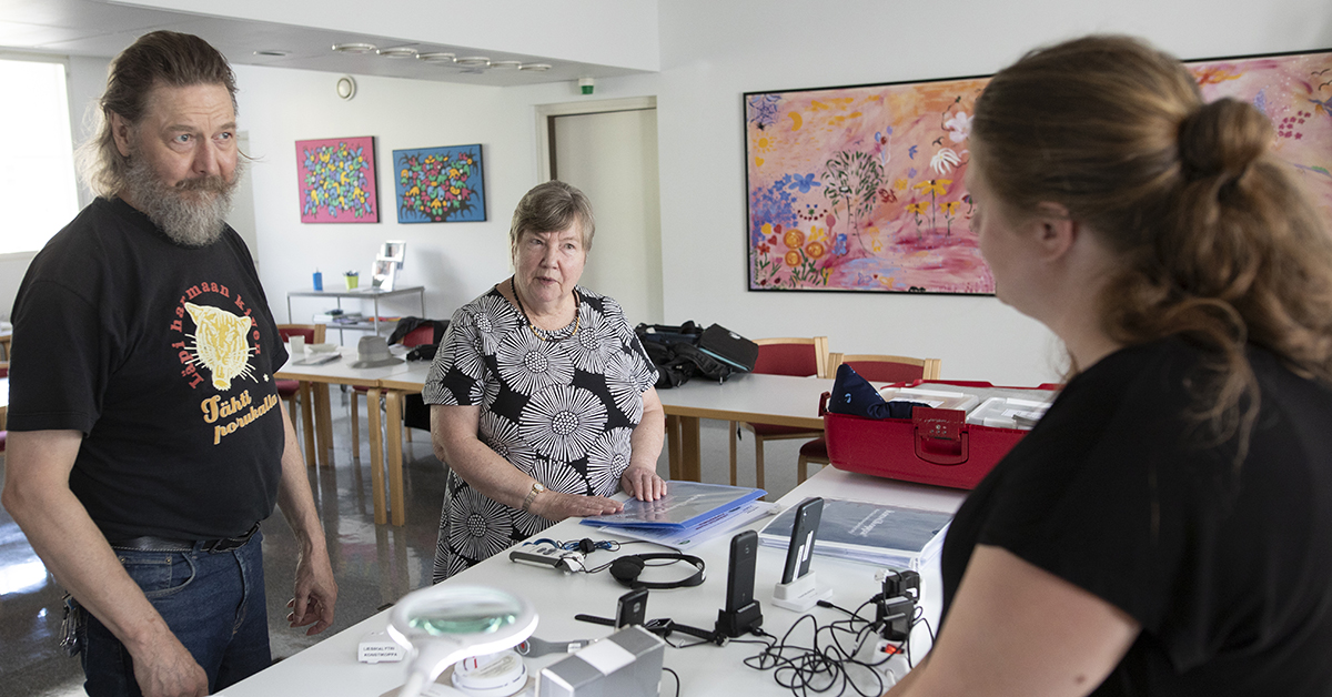 Kolme ihmistä tutkii pöydällä olevia erilaisia apuvälineitä ja laitteita.