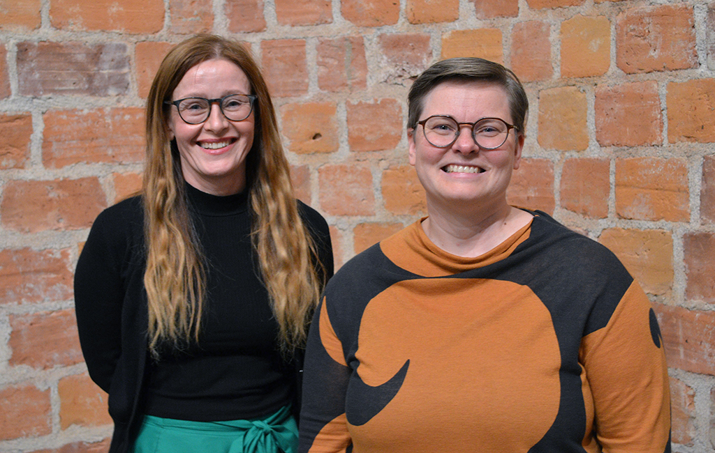 Hymyilevät Asta Leppä ja Anna Rajavuori nojaavat tiiliseinään.